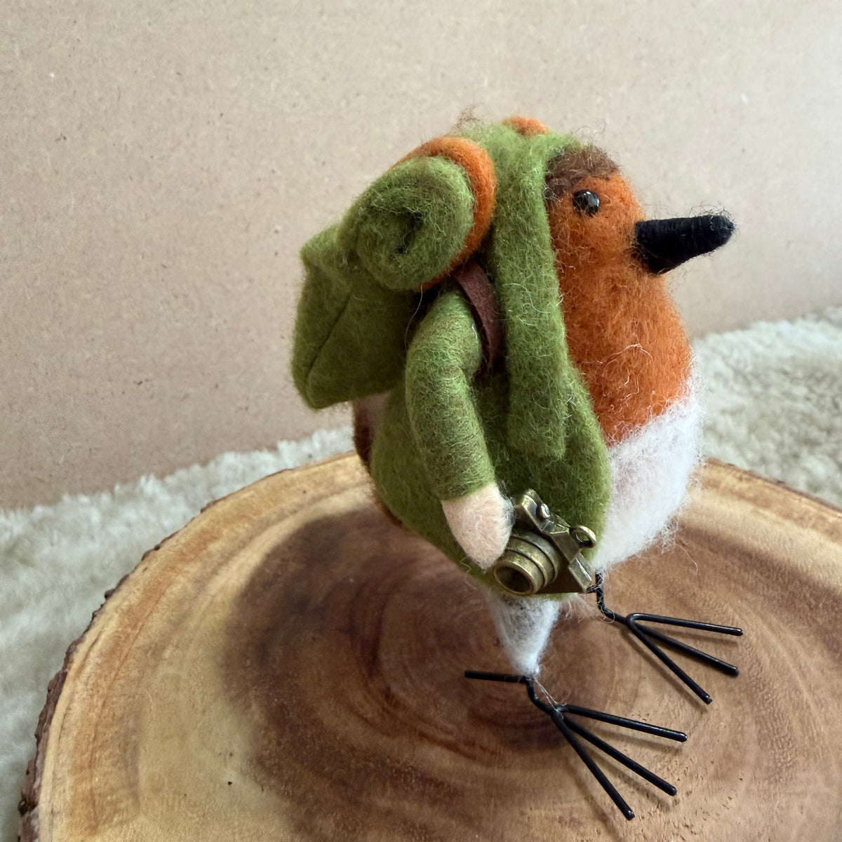 A handcrafted felted wool robin bird figurine wearing a green and orange backpack, standing on a rustic wood slice.
